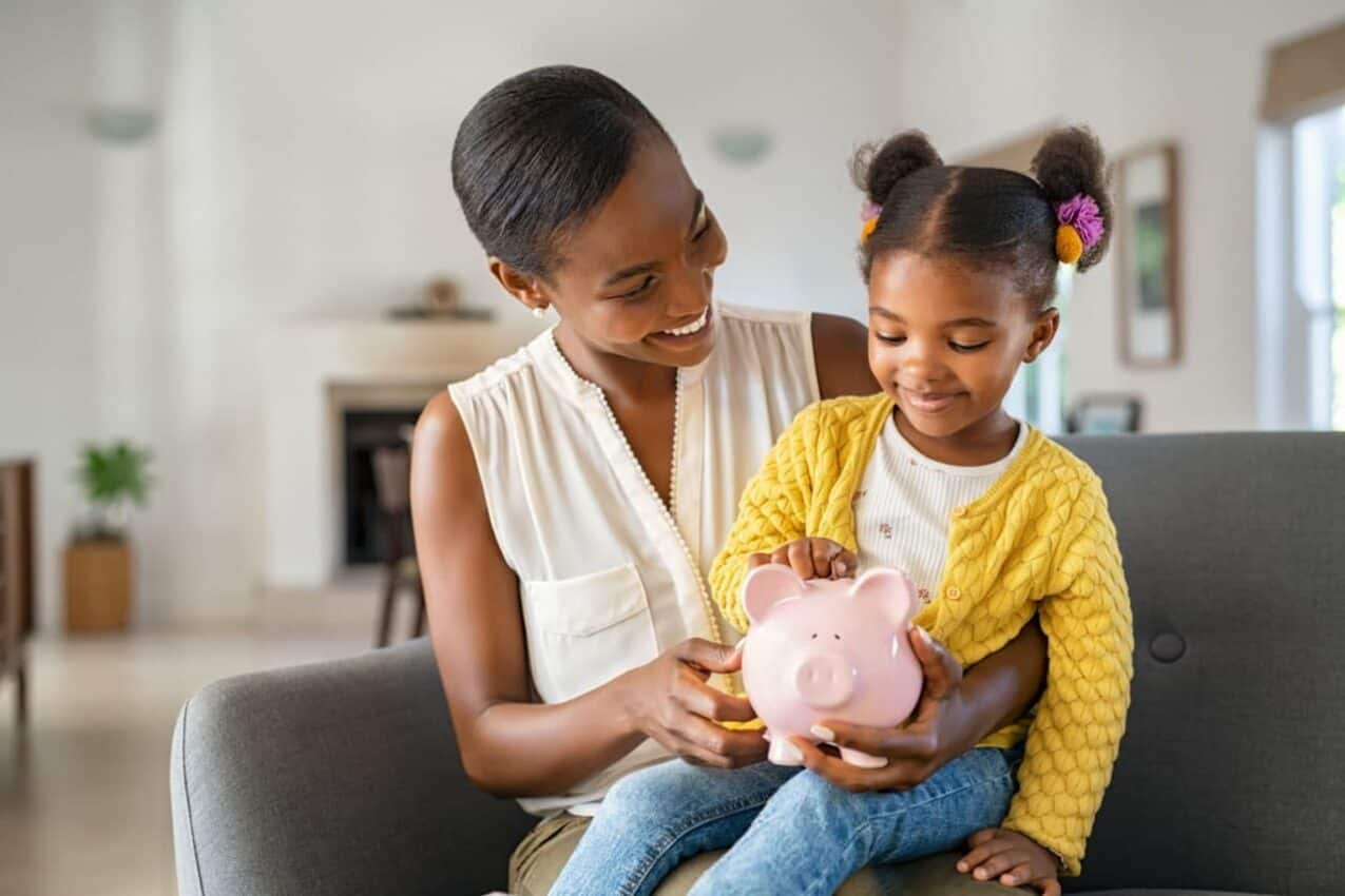 Mãe sorridente com filha pequena no colo. As duas seguram um cofrinho em formato de porco.