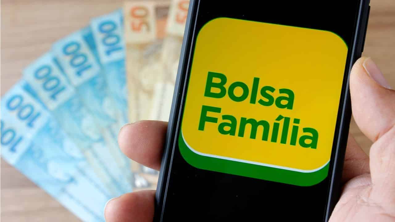 Mão segurando um celular, na tela do celular está escrito "Bolsa Família", na cor verde e o fundo amarelo. Atrás do celular, em cima de uma mesa, tem notas de 50 e 100 reais