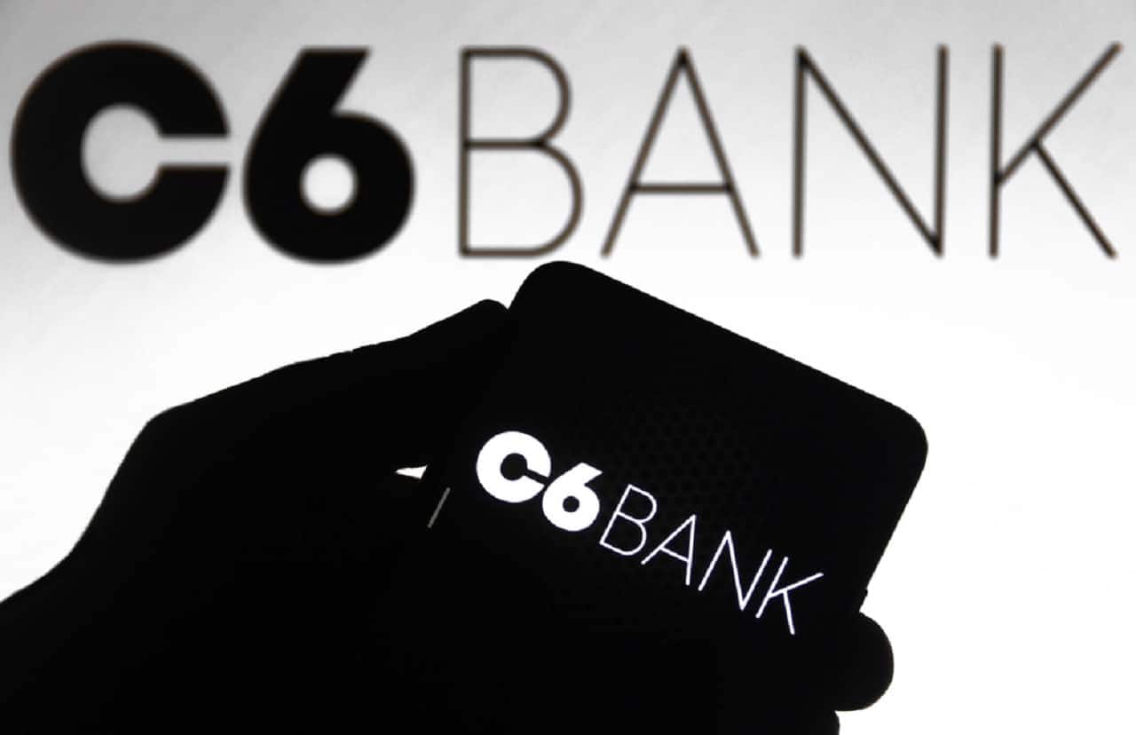 Imagem de uma mão segurando um celular com o aplicativo do c6 Bank. Ao fundo, está também a logo do C6 Bank.