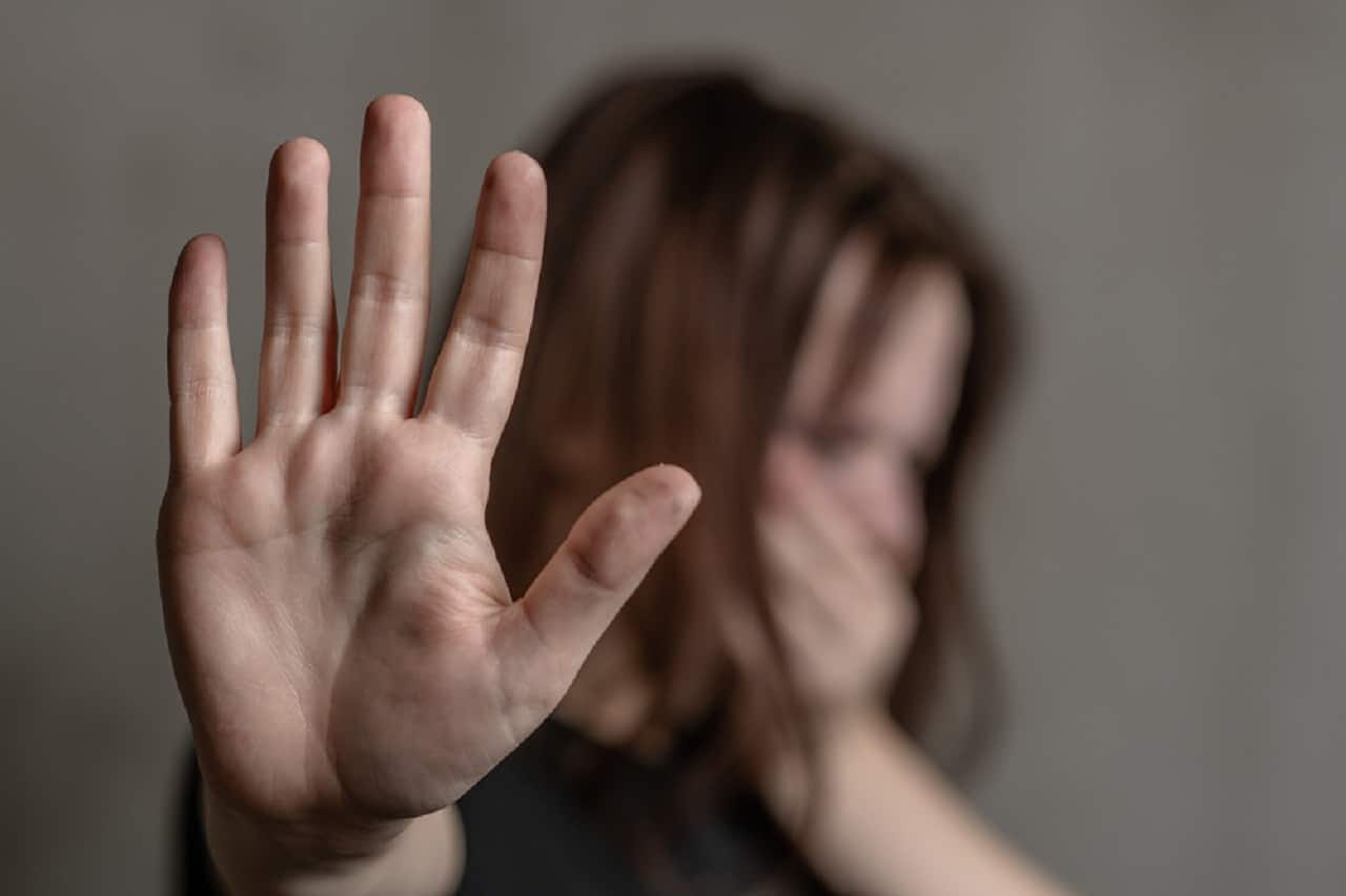 Imagem de uma mulher desfocada ao fundo tampando a própria boca com uma mão. A outra mão está nítida em primeiro plano, aberta, como se pedisse para parar.