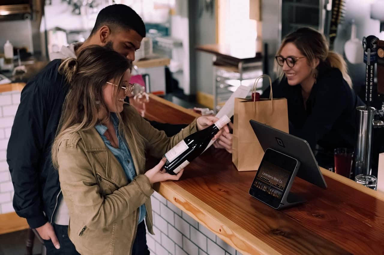 um homem negro e uma mulher branca olhando um vinho pra comprar em uma loja, sendo atendidos por uma mulher branca.