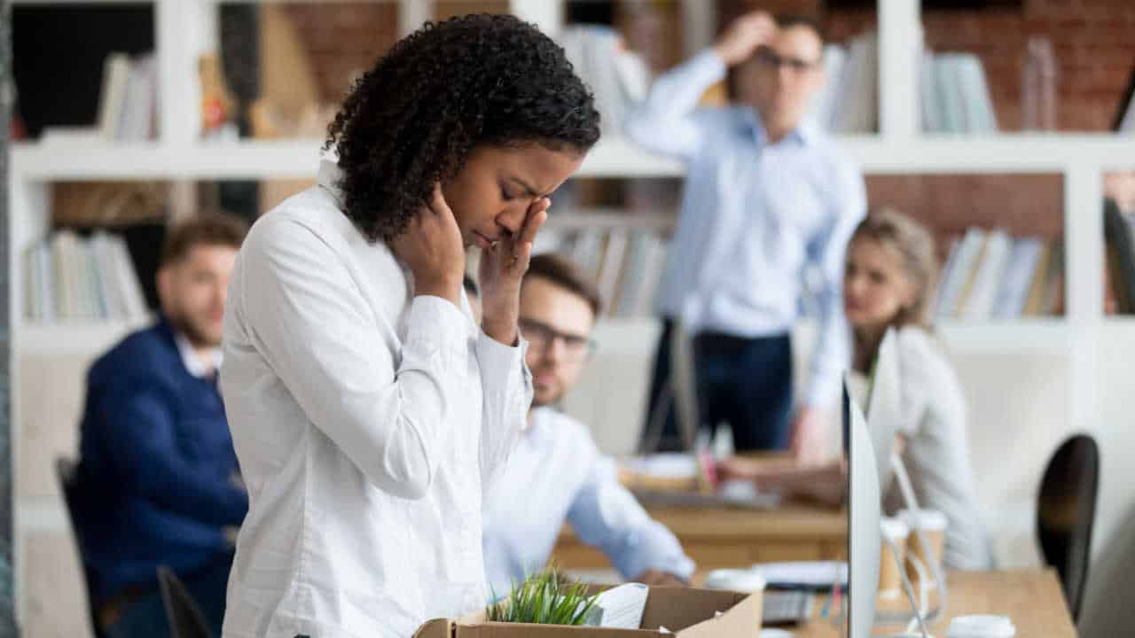 Mulher chorando depois de ser demitida da empresa. Ela está empacotando os seus pertences no escritório enquanto os outros funcionários a observam.