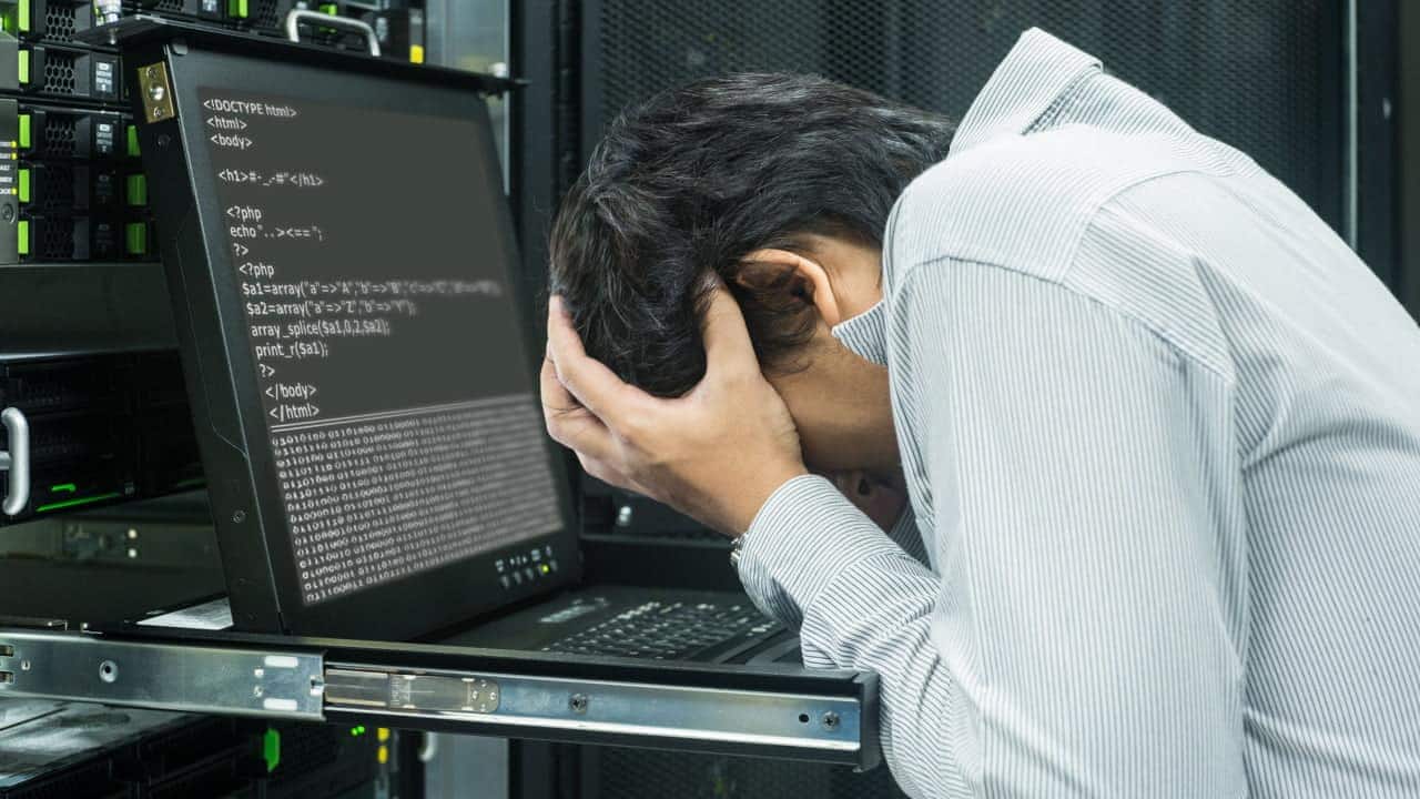 Profissional de tecnologia da informação com o rosto escondido entre as mãos diante de um computador de programação aberto