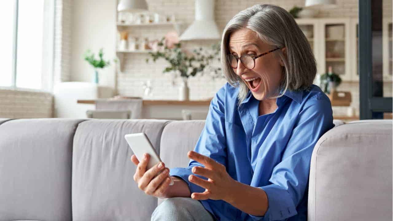 Sentada em um sofá, mulher apresenta expressão com um misto felicidade e surpresa enquanto olha para o celular