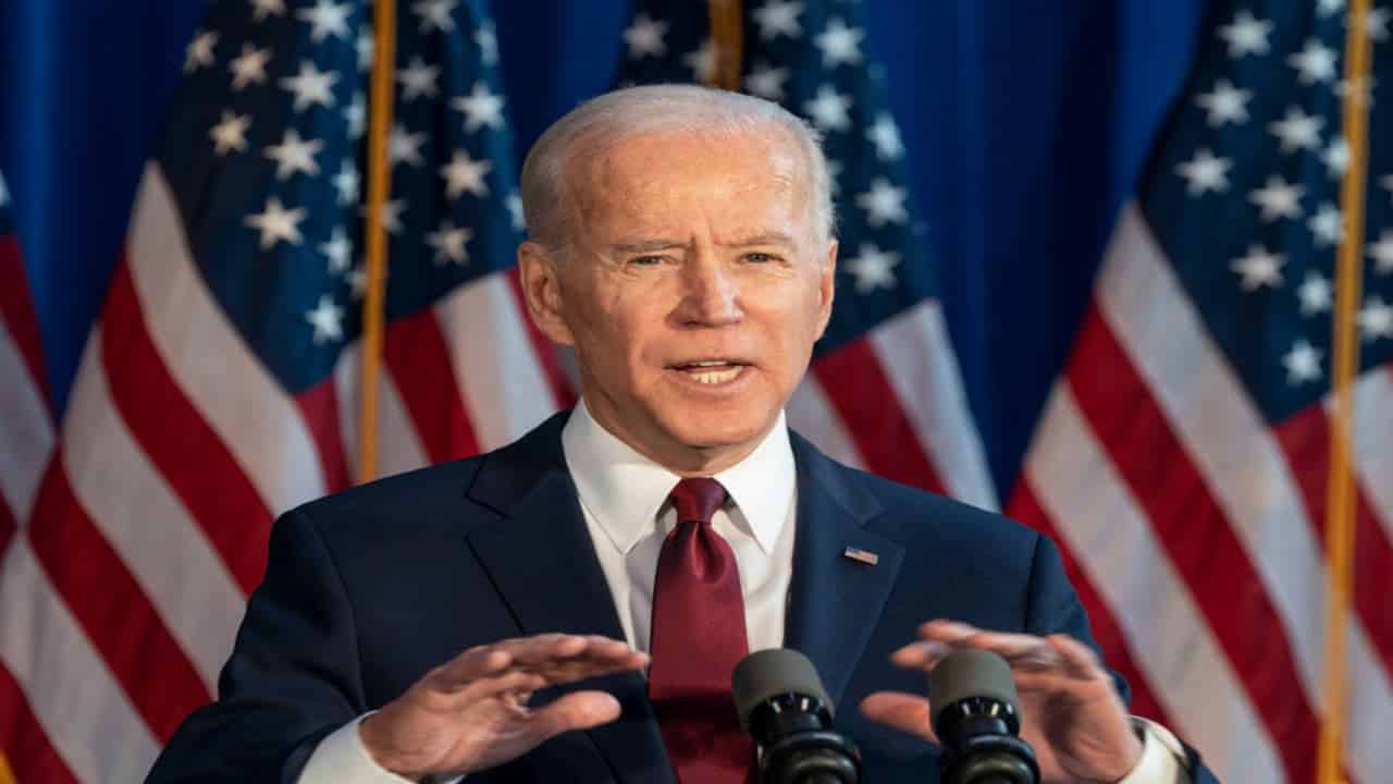 Microfones posicionados em palanque enquanto presidente dos Estados Unidos, Joe Biden, fala. Ao fundo, três bandeiras dos EUA desfocadas