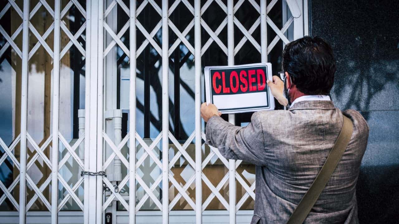 Homem de terno cinza pendurando placa com a palavra closed (fechado, em inglês) em uma portão