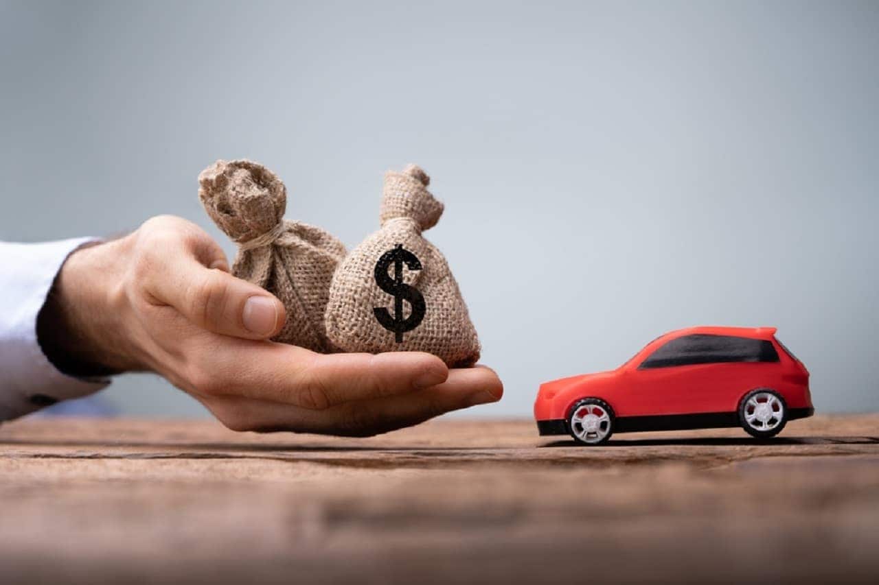 Mãe segurando dois sacos em miniatura com o símbilo "%" ao lado de um carro em miniatura.