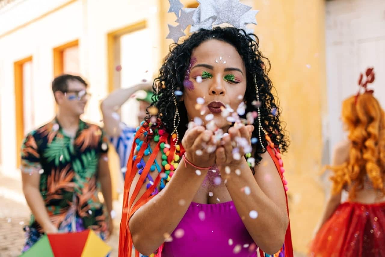 Mulher aproveitando o Carnaval ao ar livre, soprando confetes entre as duas mãos.