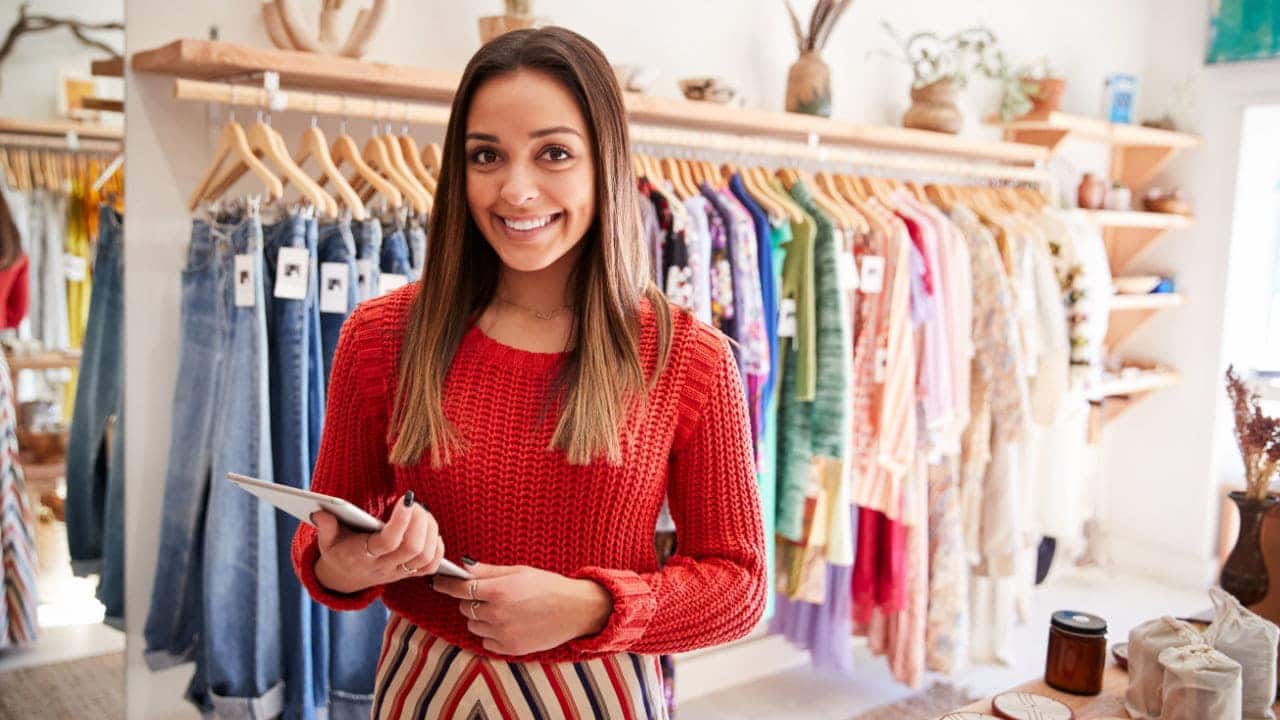 Mulher sorridente segurando um tablet no interior de uma loja, em frente a uma arara de roupas