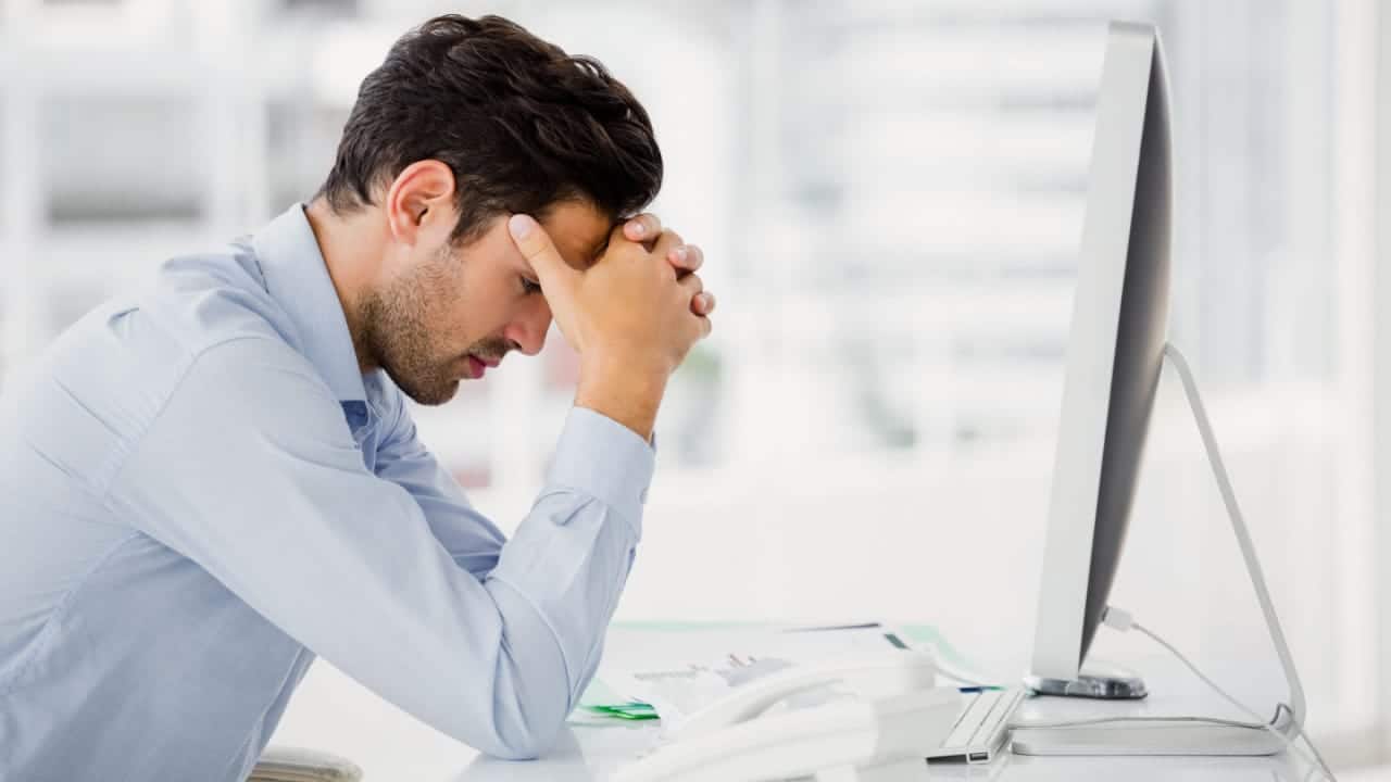 Homem de roupa social sentado em uma mesa de escritório com a cabeça apoiada nas duas mãos, aparentando frustração. À sua frente, há uma tela de computador.