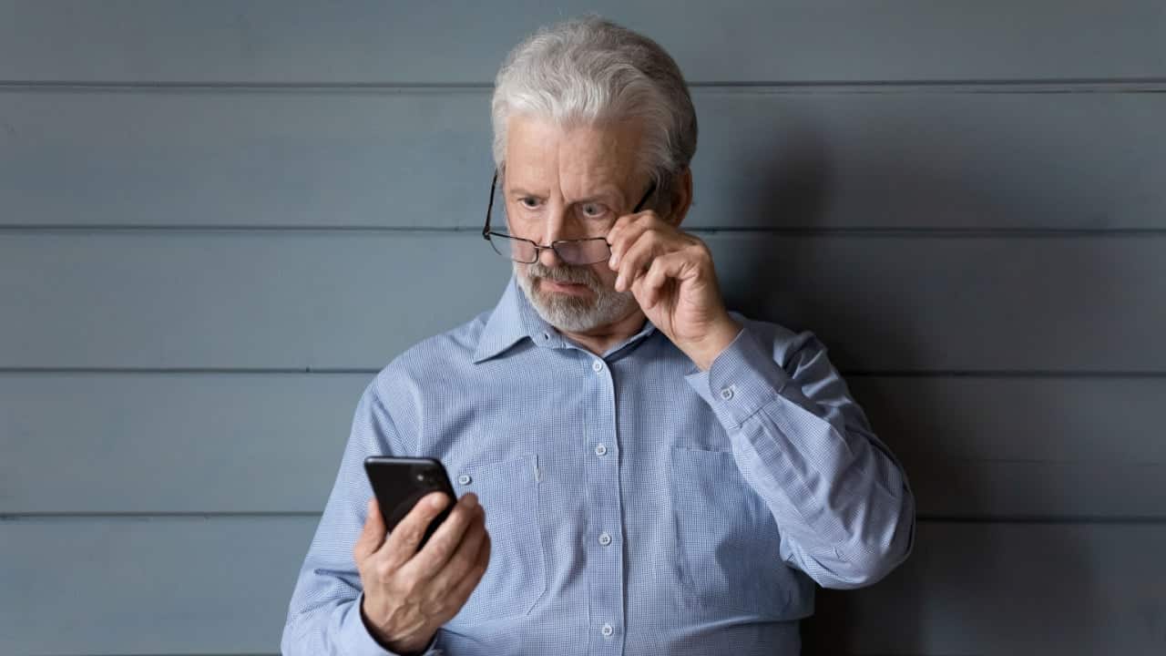 Homem idoso segurando os óculos no rosto e olhando para um celular com expressão de choque