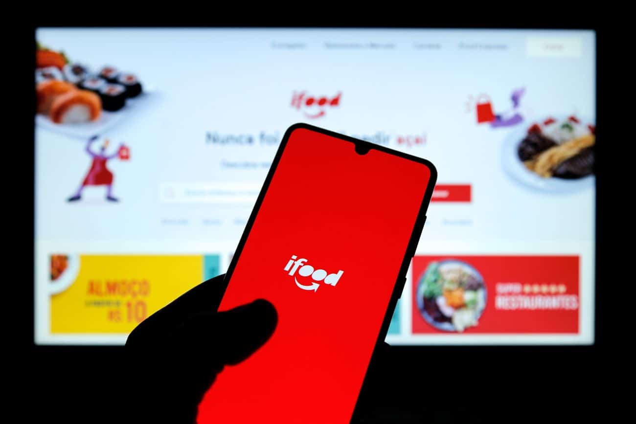 Sombra de uma mão segurando um celular que mostra na tela o logo do aplicativo iFood em fundo vermelho