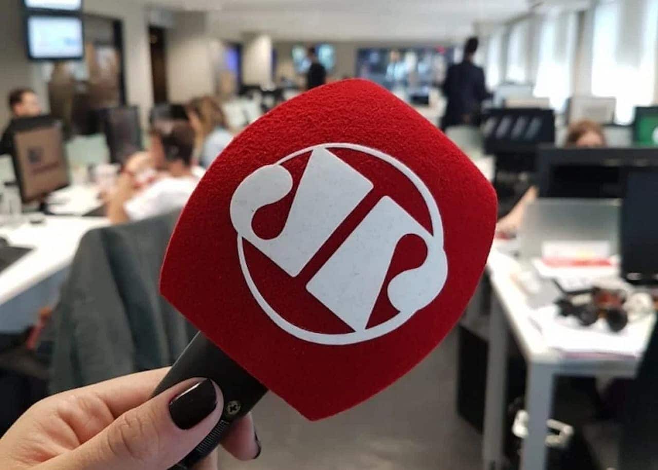 mão feminina segurando microfone vermelho da Jovem Pan. Ao fundo da imagem a redação da empresa