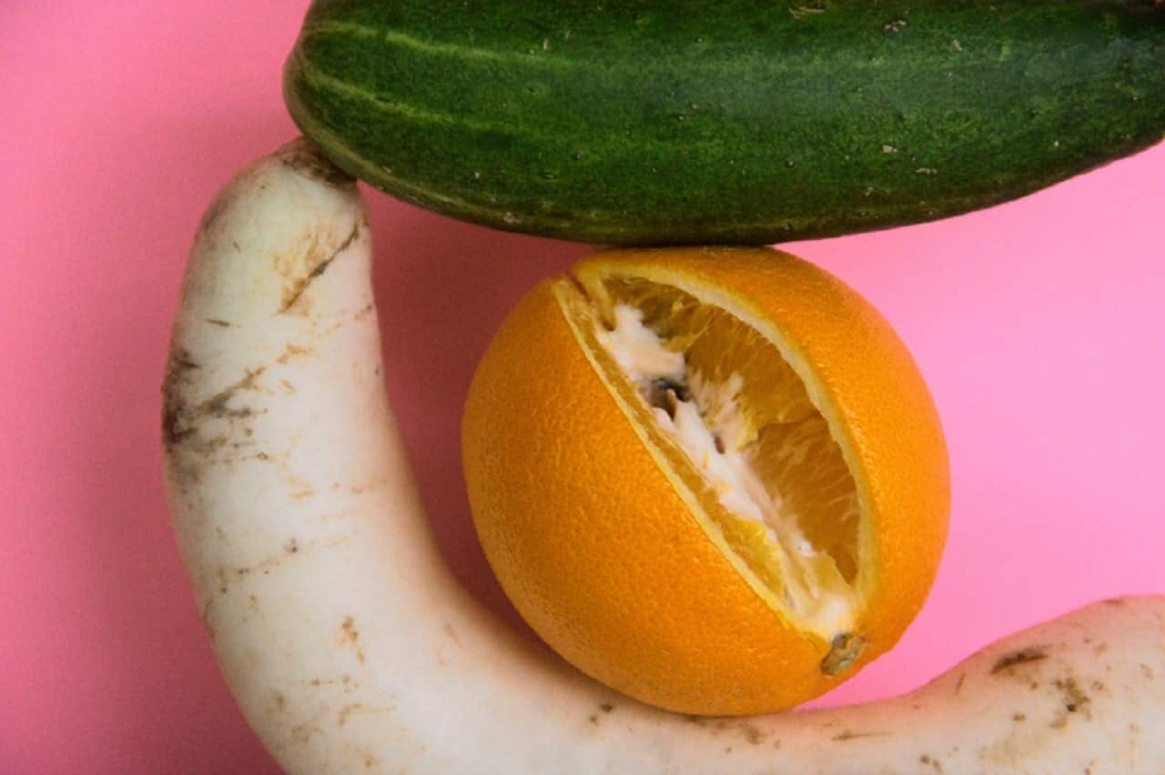 Imagem de um pepino, uma laranja cortada e uma mandioca em um fundo rosa.
