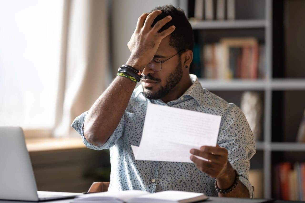 Homem com a mão na cabeça e semblante de preocupação segurando um pedaço de papel com uma multa, ou seja, uma advertência financeira