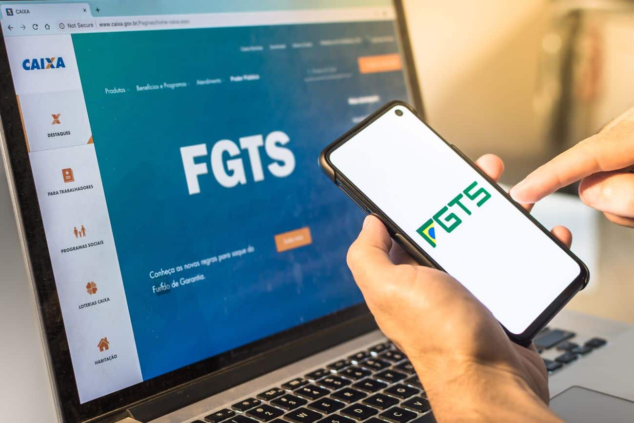 Mão masculina segurando celular com a logo do FGTS. No fundo, notebook mostrando tela inicial do site do FGTS