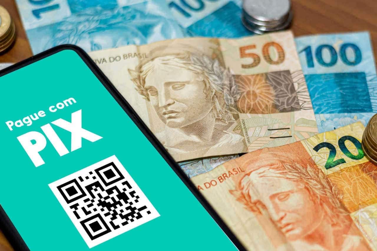 Tela do celular exibindo os dizeres "pague com pix" e algumas notas de dinheiro próximas ao aparelho