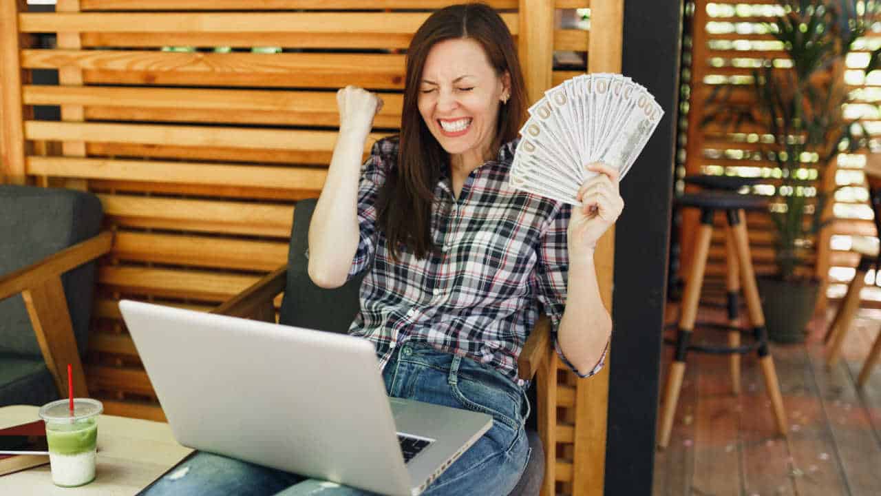 Mulher feliz com notebook no colo e um “leque” de dinheiro na mão