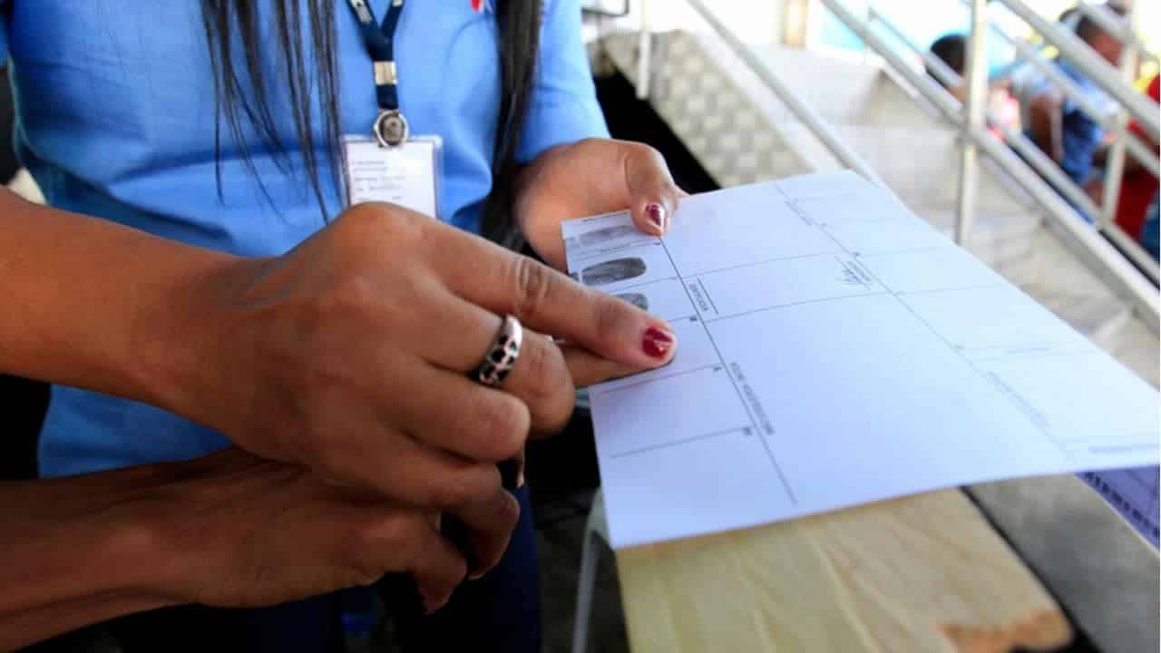 Pessoa tendo impressão digital colhida por outra para emissão de RG