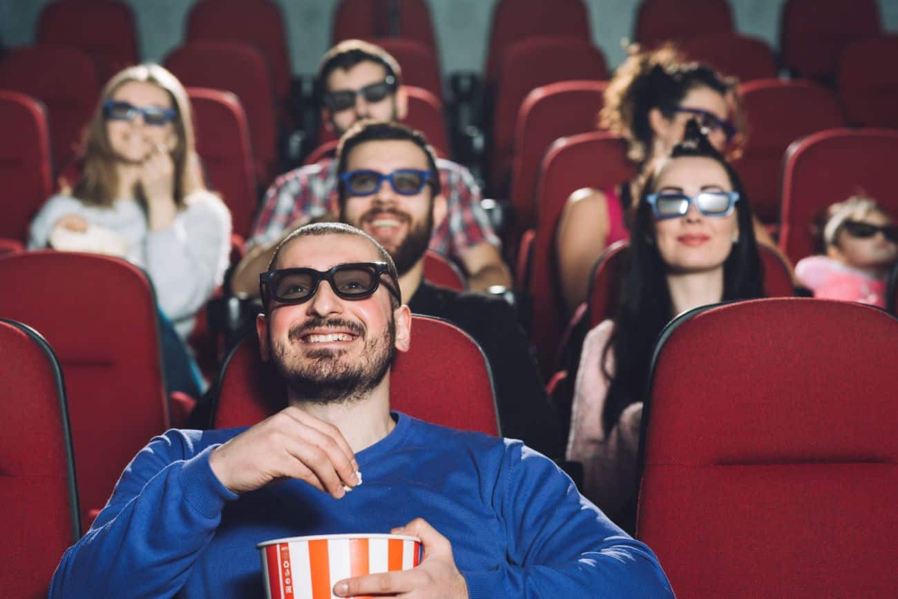 Diversas pessoas sentadas em uma sala de cinema usando óculos 3D e comendo pipoca