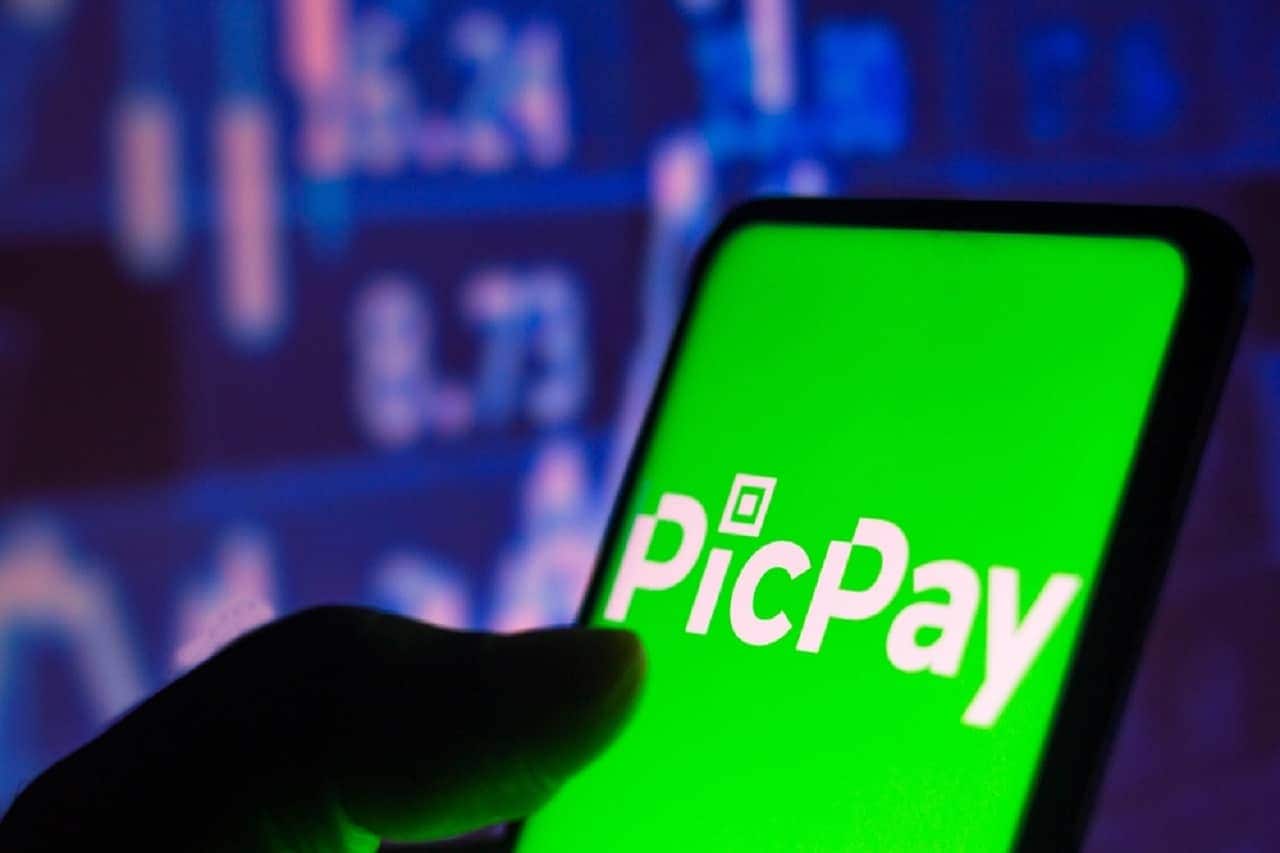 Imagem de uma mão segurando um celular com a tela do aplicativo do PicPay aberto. Ao fundo, computadores desfocados.