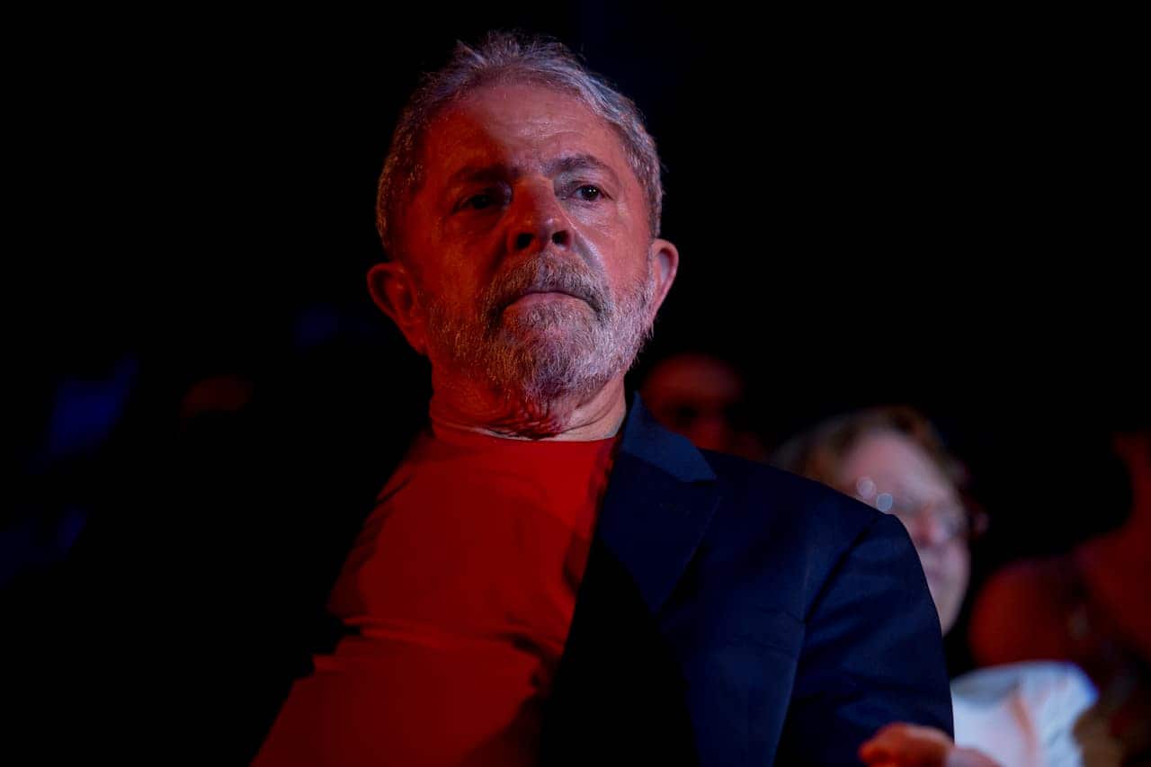 Presidente Lula sentado em um lugar um pouco escuro usando uma blusa vermelha por baixo de uma camisa azul