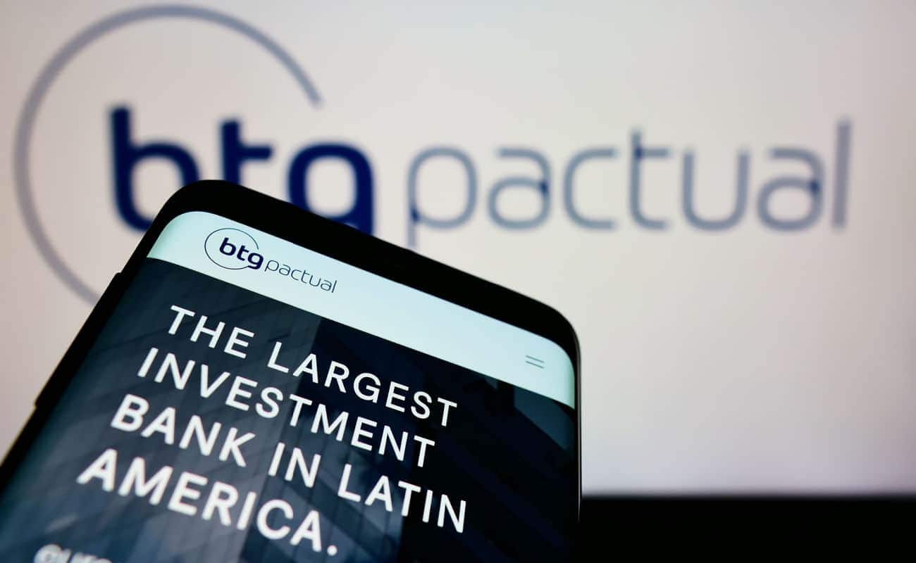 No fundo está a logo do BTG Pactual, na frete um celeular com o site da empresa aberto, onde se lê "O maior banco de investimento da América Larina" em inglês
