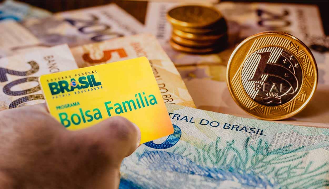 Mão segurando um cartão do Bolsa Família com cédulas e moedas de real ao fundo