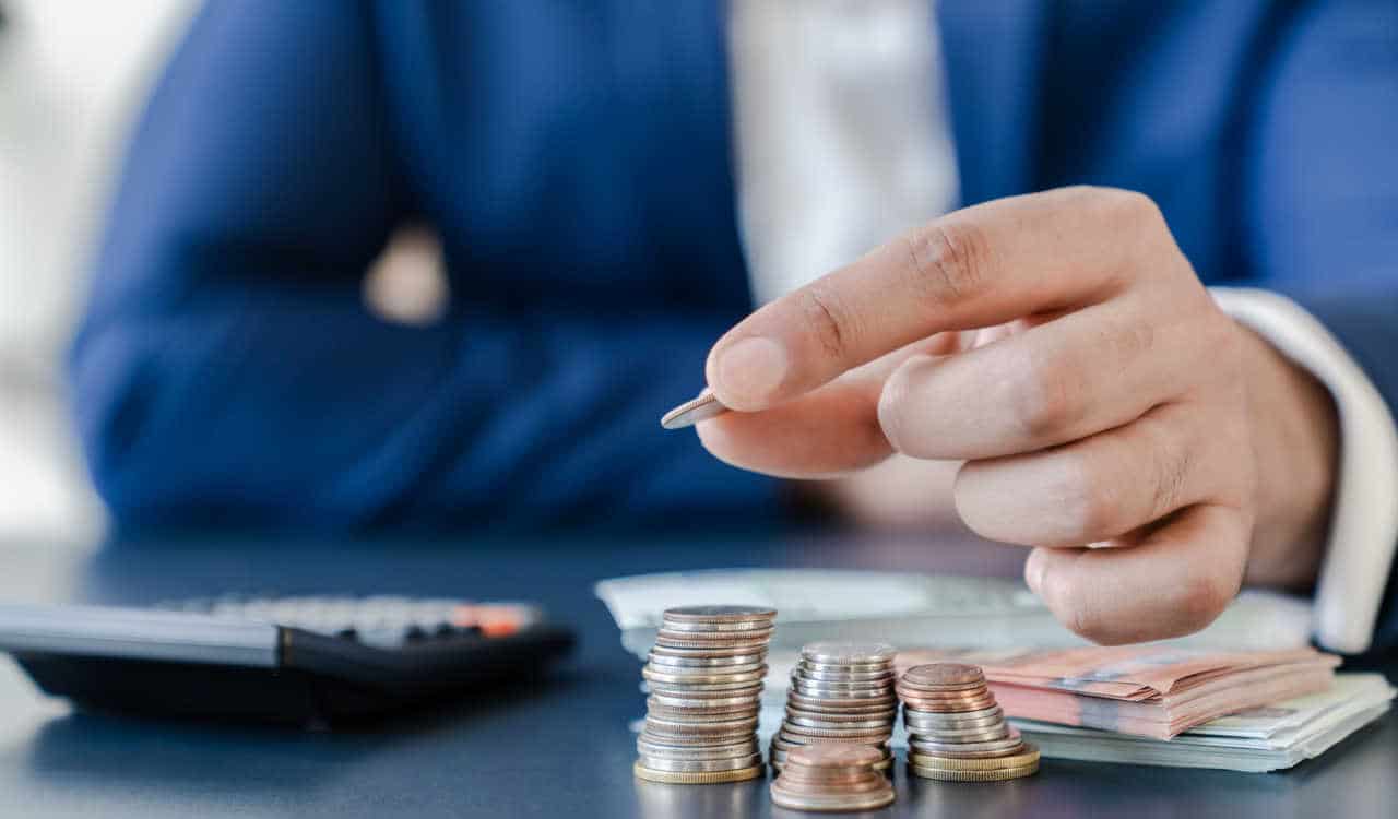 Homem vestido de terno empilhando moedas na frente de notas de dinheiro e ao lado de uma calculadora