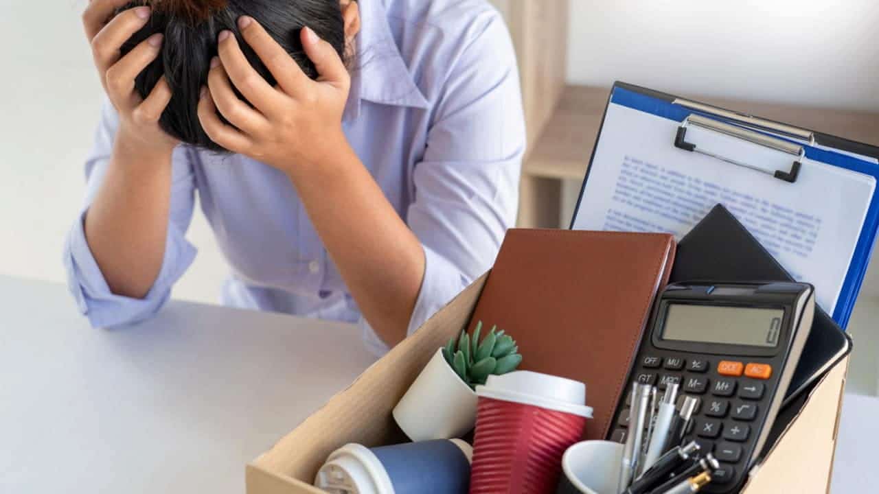Homem sentado à mesa de um escritório com a cabeça apoiada nas mãos após perder o emprego, ao lado dele tem uma caixa com pertences pessoais.