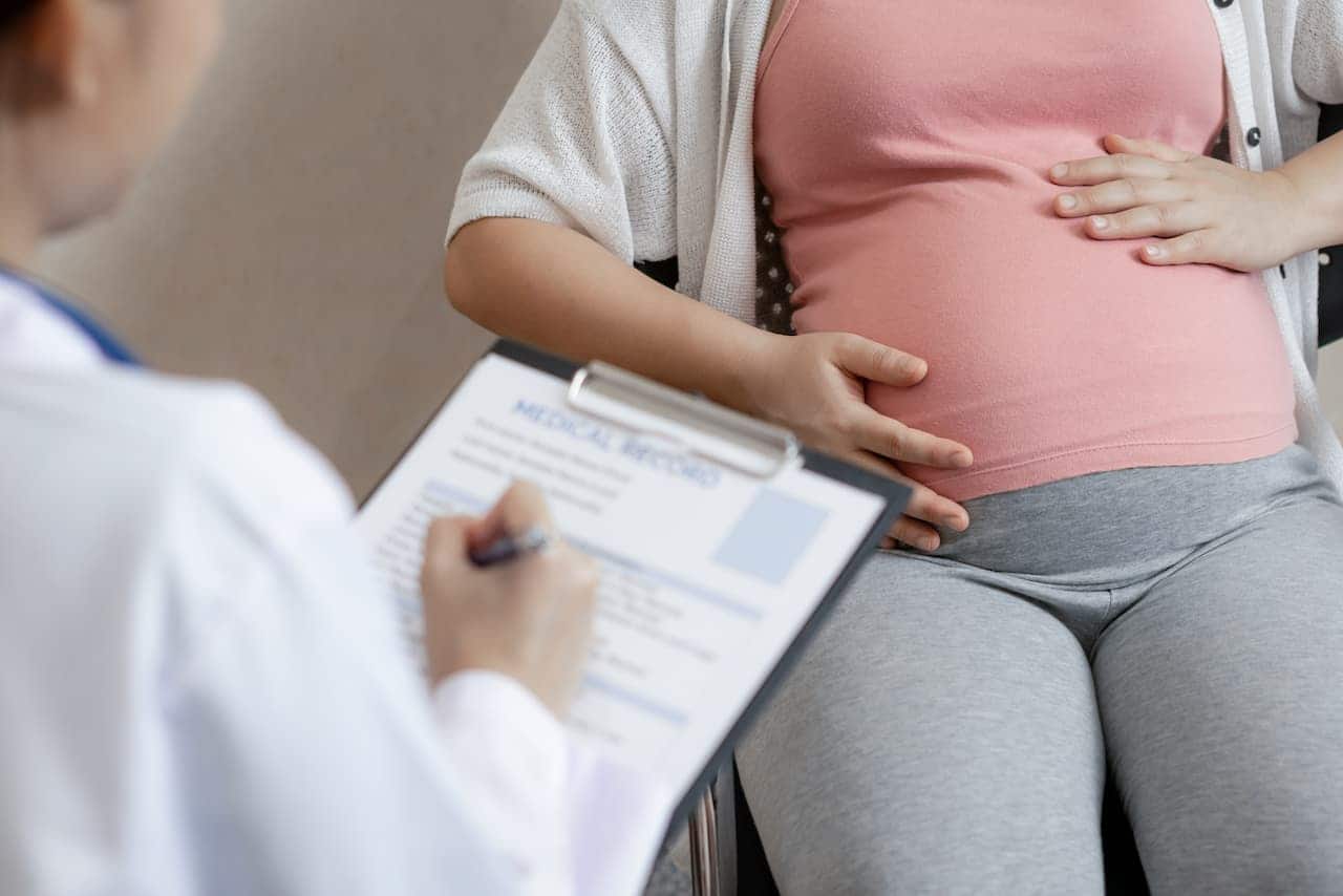 Mulher grávida com a mão na barriga sentada de frente para uma médica que está com papéis e caneta nas mãos.
