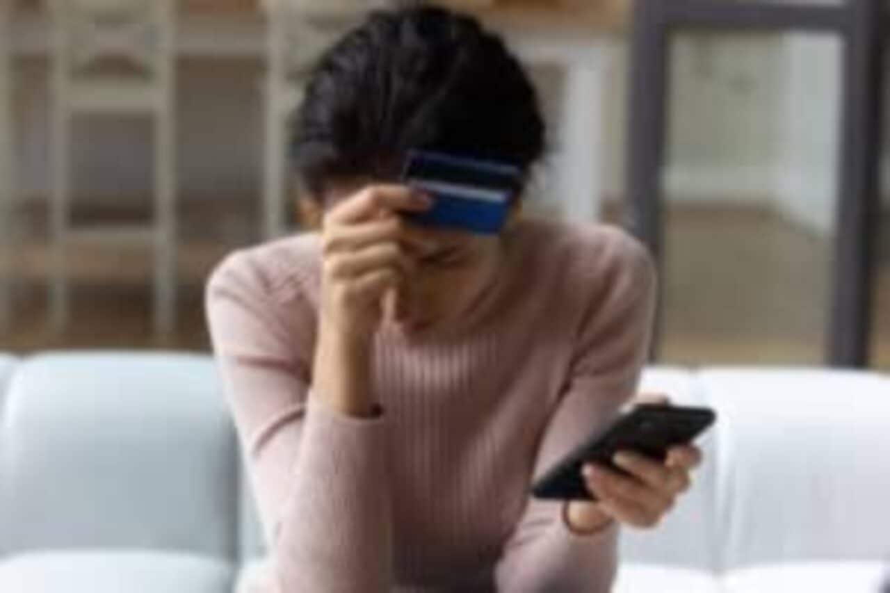Mulher sentada em sofá com a cabeça baixa segurando um cartão e um celular