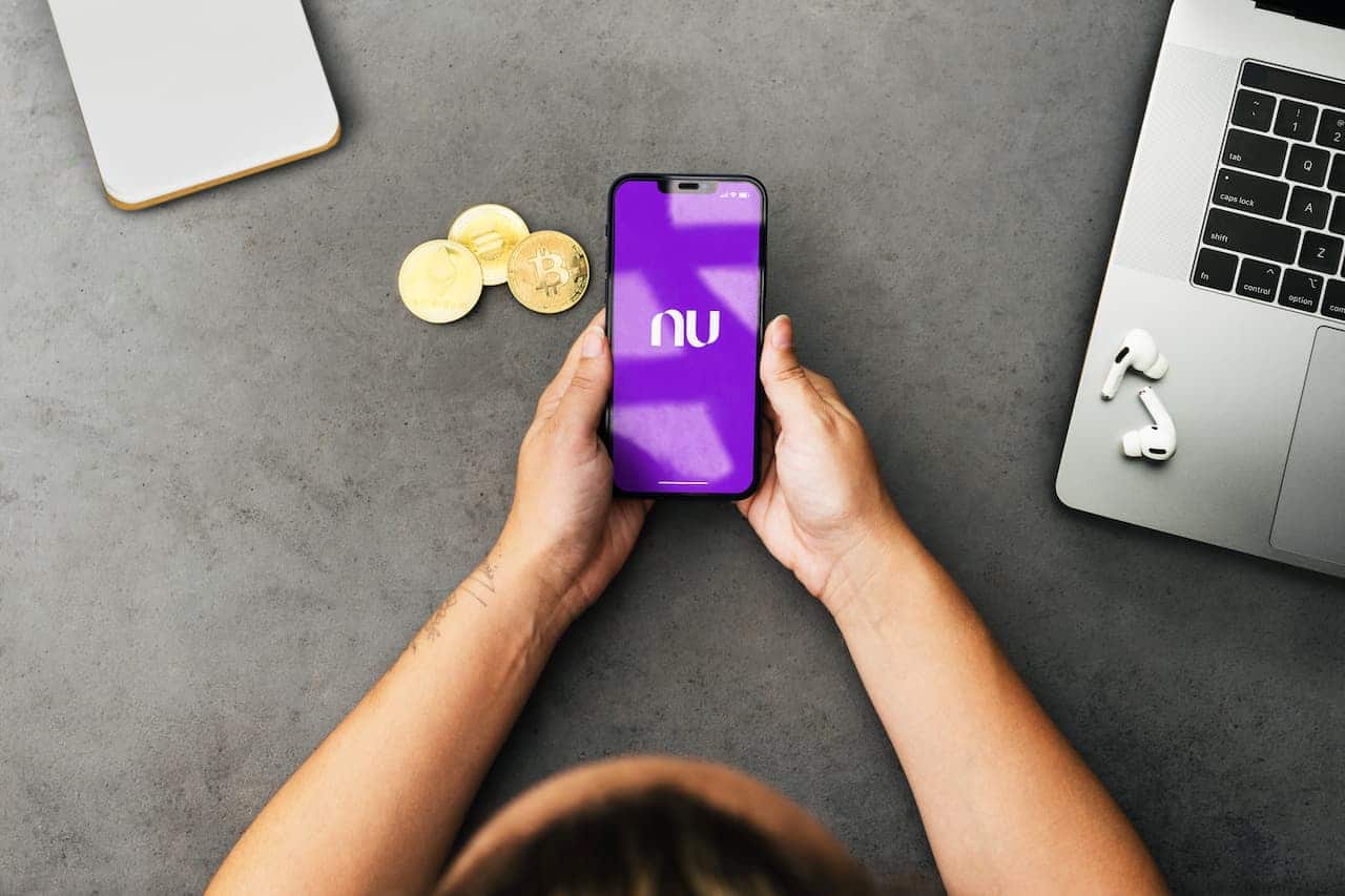 Imagem de pessoa segurando celular com app Nubank aberto e criptomoedas em cima da mesa