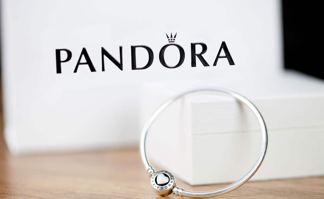 Em primeiro plano aparece um bracelete de prata. Ao fundo pode ser visto a logo da Pandora