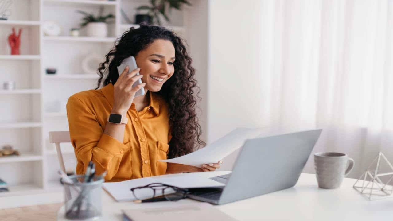 Mulher sentada em frente a uma mesa de trabalho olhando para um notebook e segurando um smartphone próximo ao ouvido