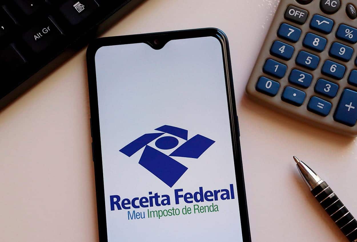 Celular com aplicativo do Imposto de Renda aberto ao lado de calculadora e caneta