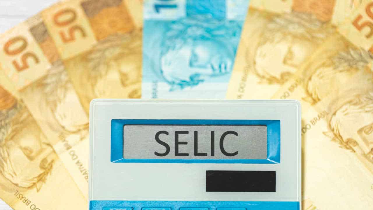 a palavra “Selic” escrita no visor de uma calculadora que está em cima de notas de R$ 50 e R$ 100