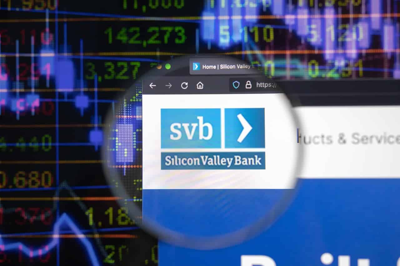 Logo do banco SVB em destaque na tela de um computador. Ao fundo gráficos da bolsa de valores