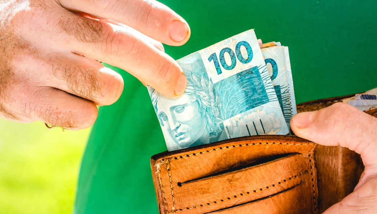 Mãos de um homem guardando dinheiro, com notas de R$ 100 em evidência, na carteira.
