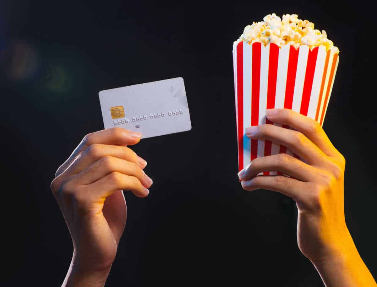 uma mão segurando um cartão de crédito e outra segurando um saco de pipoca de cinema, o fundo é preto dando destaque apenas às mãos e aos objetos