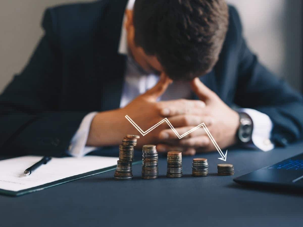 Imagem de empresário triste, na frente dele moedas empilhadas em forma de gráfico com uma seta indicando para baixo, representando o prejuízo do seu negócio.
