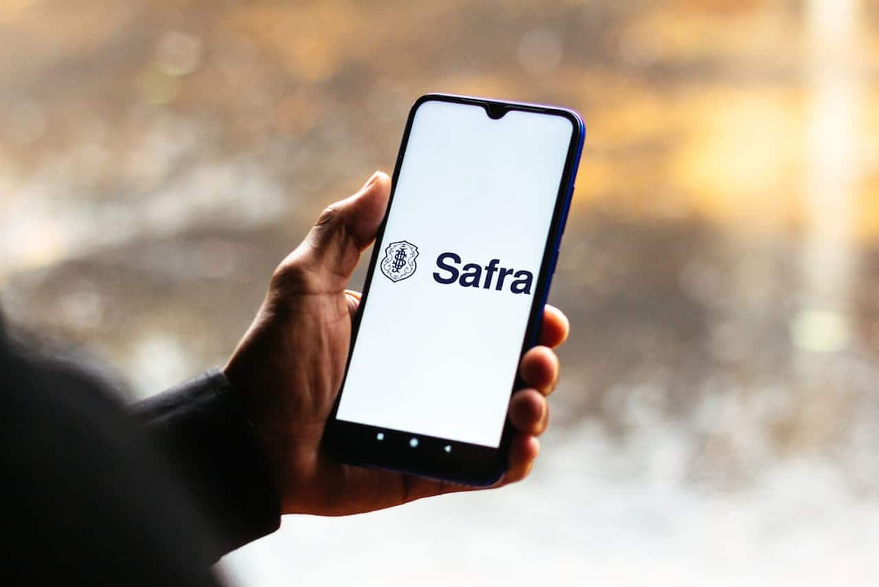 Mão segurando celular que exibe logotipo do Banco Safra. Fundo desfocado