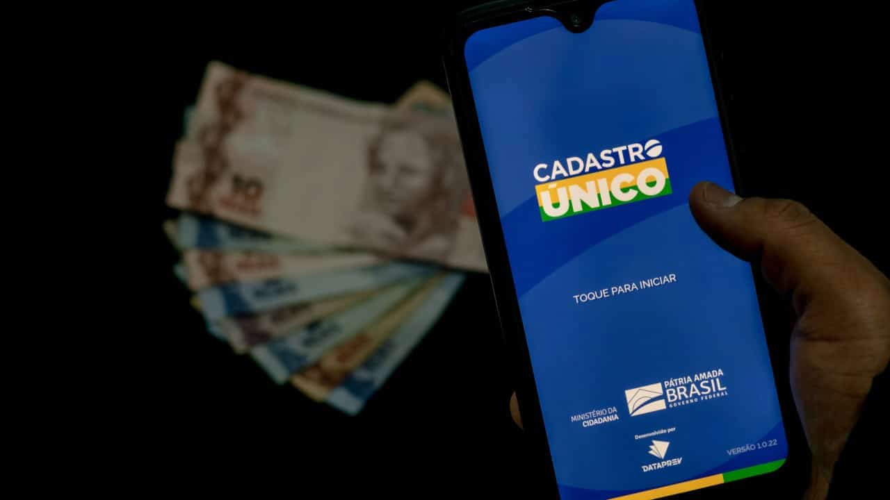 Mão acessa o app do Cadastro Único em um smartphone e também é possível ver algumas notas de dinheiro ao fundo