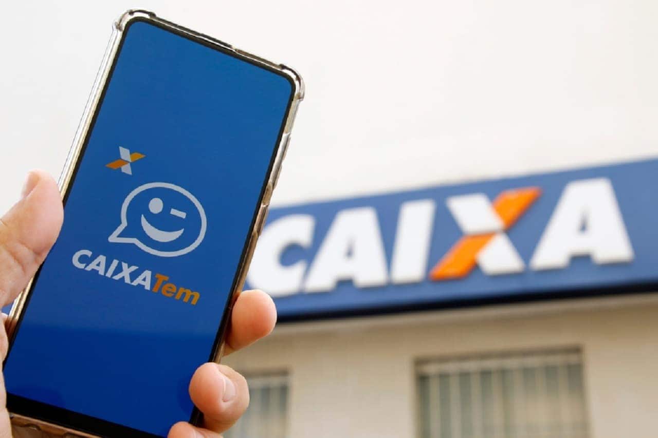 Imagem de uma mão segurando um celular com o aplicativo Caixa Tem aberto. Ao fundo, uma agência da Caixa Econômica Federal.