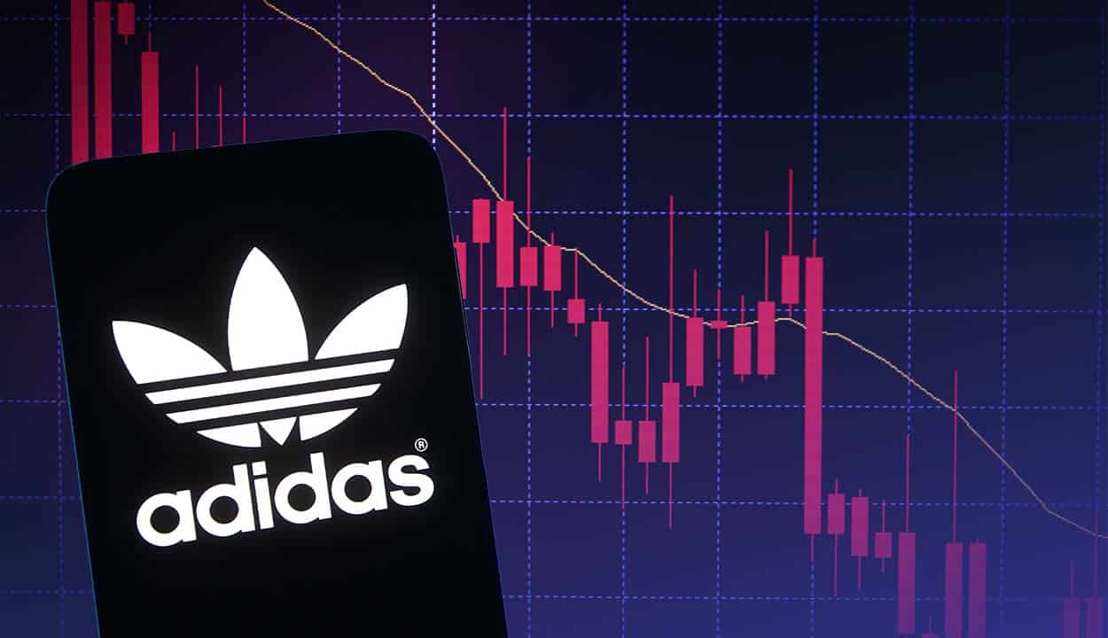 Celular com logo da Adidas aberto e tela com gráficos em queda ao fundo