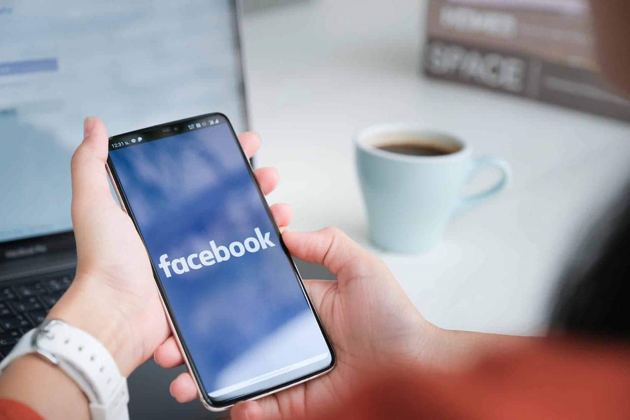 Mãos segurando um celular que mostra na tela a logo do Facebook em um fundo azul