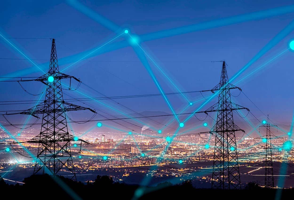 Torres de energia elétrica, interligadas por fios. Ao fundo cidade iluminada.