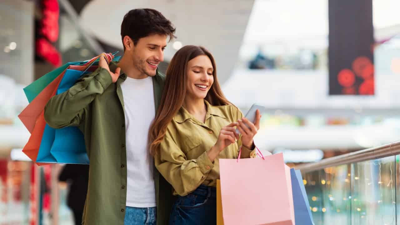 casal feliz com sacolas de compras olhando para um celular, enquanto viajam