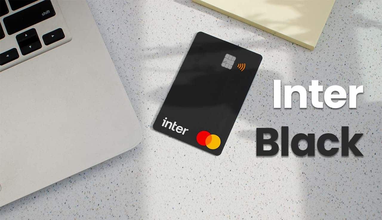 Cartão black do Banco Inter sobre uma mesa cinza e ao lado um notebook cinza também sobre a mesa, ao lado do cartão está escrito Inter Black