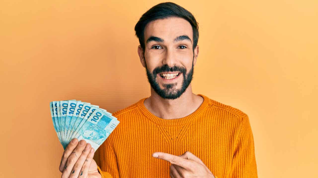 Homem segurando notas de R$ 100 em uma mão, com a outra mão ele aponta para o dinheiro e sorri
